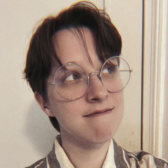 Auden Patrick, a white nonbinary person with brown hair and silver round glasses, looking to the right.